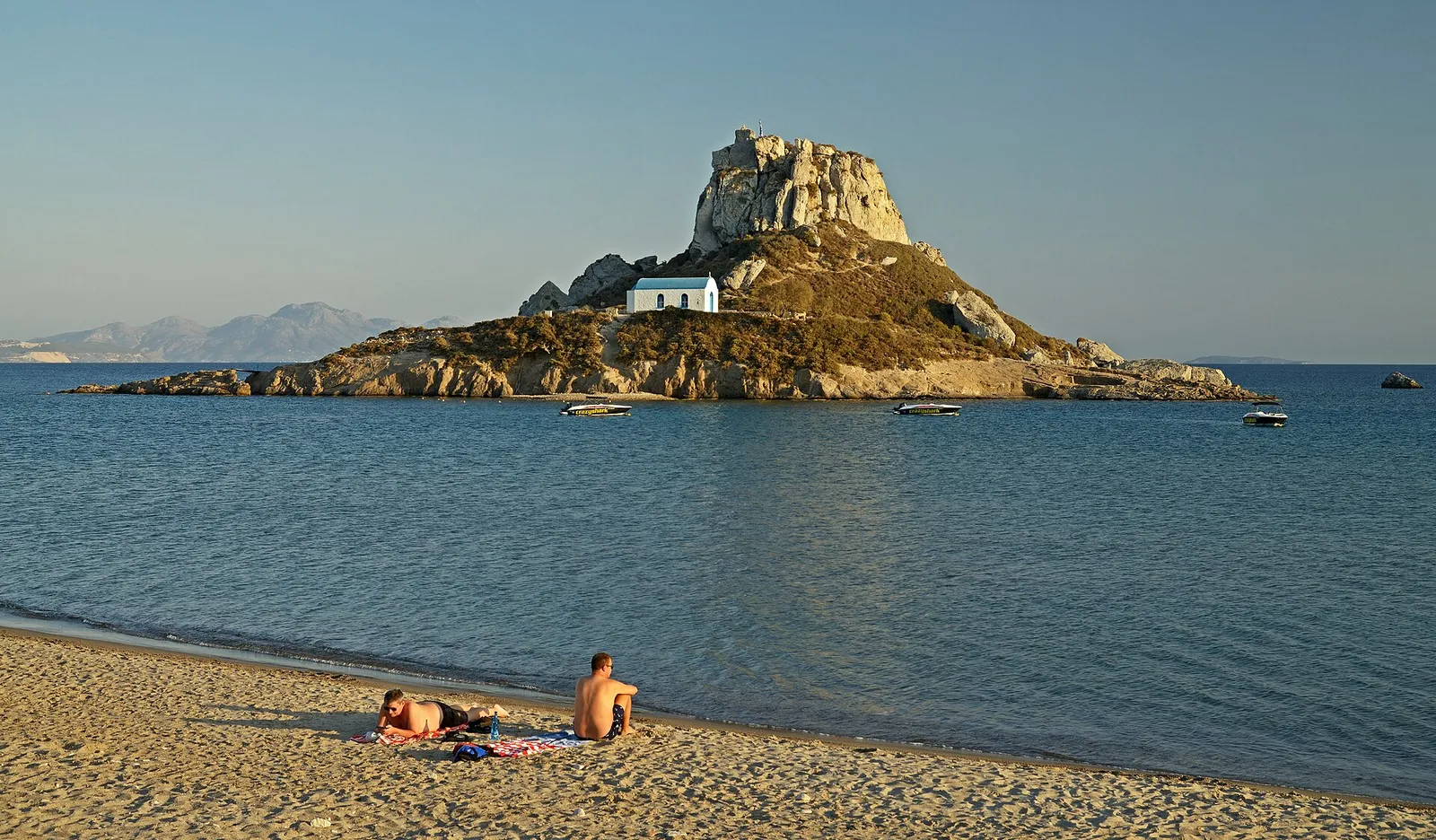 A small rocky islet crowned with a whitewashed chapel just off a sandy beach in Kos, Greece, with boats moored in calm water