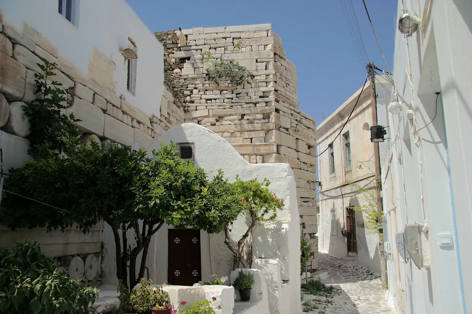 סמטה לבנה צרה עם קשתות אבן ופרחים סגולים בכפר נאוסה בפארוס, יוון