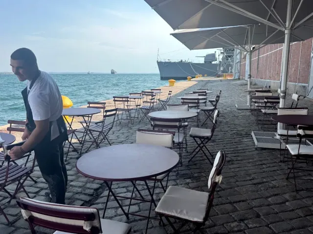 Round tables on a stone quay facing the water, and a man arranging chairs