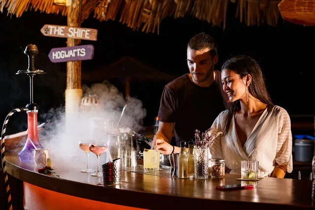 A bartender makes a smoking cocktail for a smiling woman, at a wooden counter under a straw roof