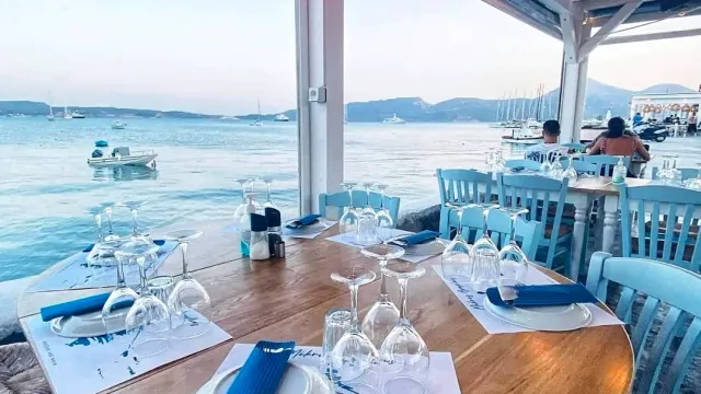 A laid table with wine glasses and pale blue chairs facing a bay with boats
