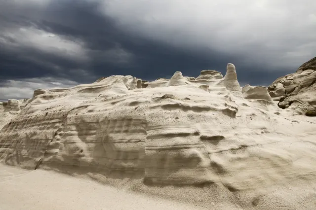 White volcanic rock carved into layers above the sand, under a cloudy sky