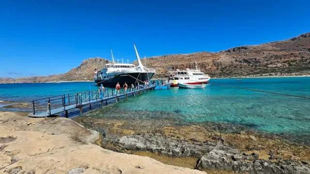 A wooden jetty over shallow turquoise water leading to two ships, and bare hills behind