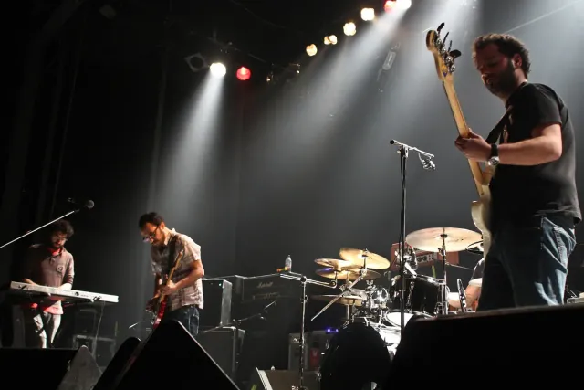 A band playing on a stage lit by spotlights, with a guitar, keyboards and drums