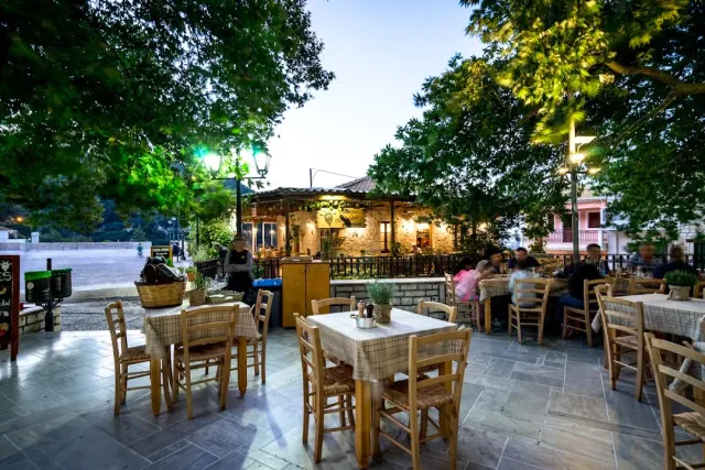 Wooden tables in a tree shaded square, facing a lit stone building in the evening