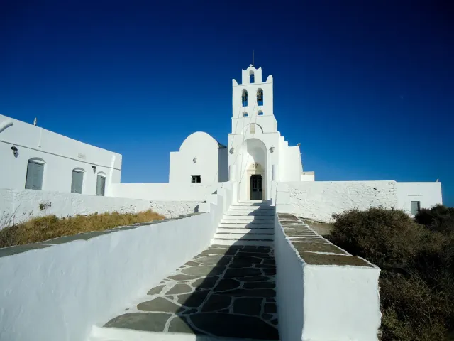 A whitewashed monastery with a bell tower at the edge of a rock, reached by a stone path