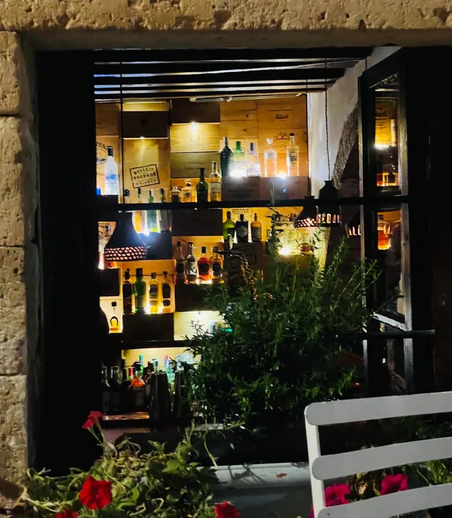 A stone framed window reveals lit bottle shelves, with plants and red flowers in front