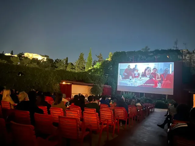 An open air cinema at night with red chairs, an audience watching and a lit screen among the trees