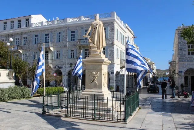 A paved square with a statue, Greek flags and a large neoclassical building