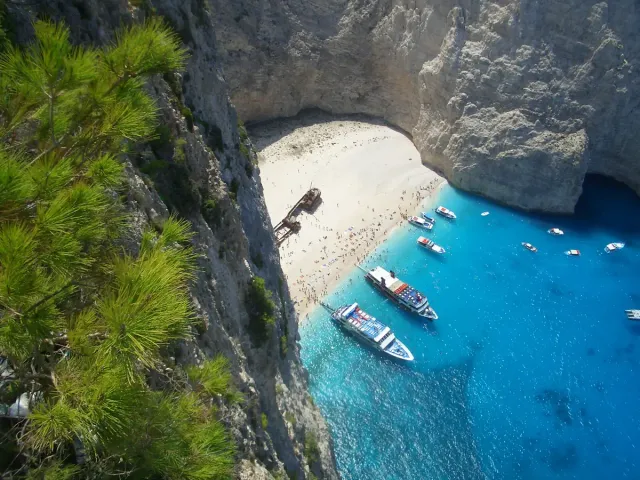 A view from above of a cove ringed by limestone cliffs, a rusted wreck on white pebbles and boats in bright blue water