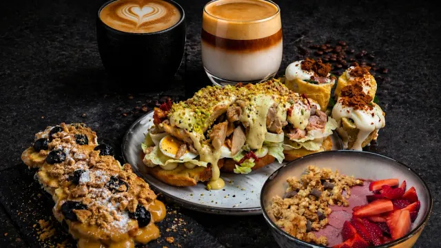 An open sandwich with chicken and sauce, a pastry, a bowl of fruit and coffee on a dark surface