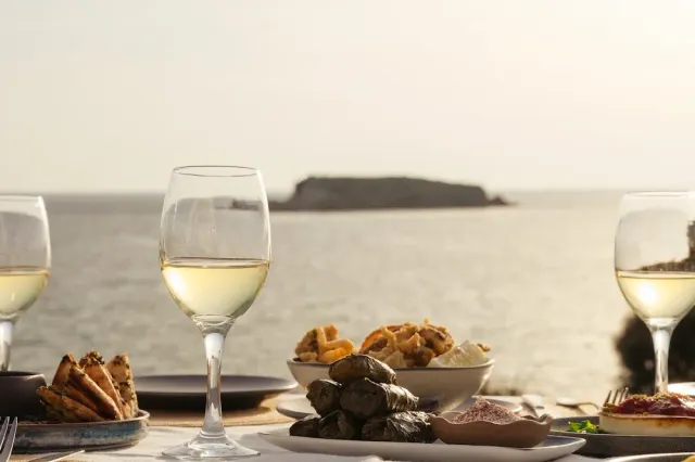 A table facing the sea with glasses of white wine, stuffed vine leaves and fritters, and an islet behind
