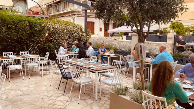 A paved courtyard with white chairs, a planted olive tree and diners at the tables