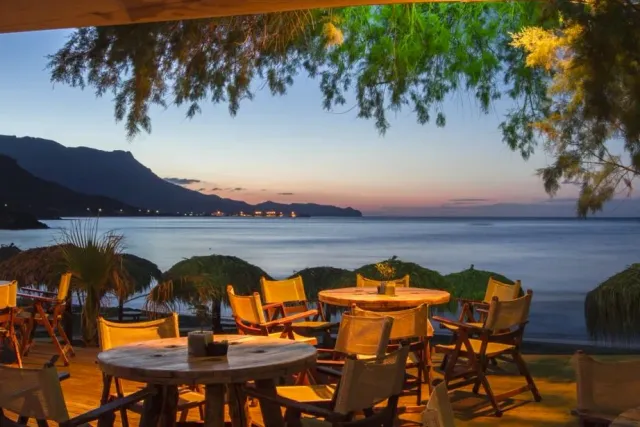 Round tables and canvas chairs under branches, facing a bay and mountains at dusk