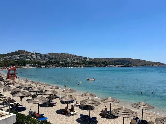 A sandy beach with straw umbrellas and sun loungers facing turquoise water and a built up hill behind