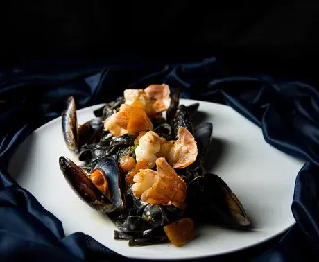 A plate of black pasta with mussels and prawns, on dark blue fabric against a dark background