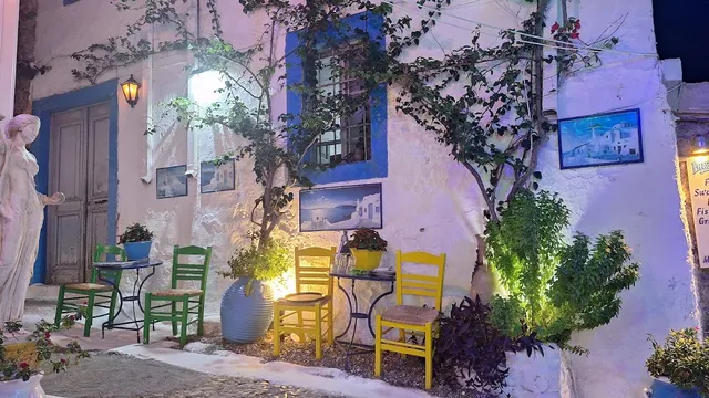 Colourful chairs and small tables before a white wall with a blue window and plants, lit in the evening