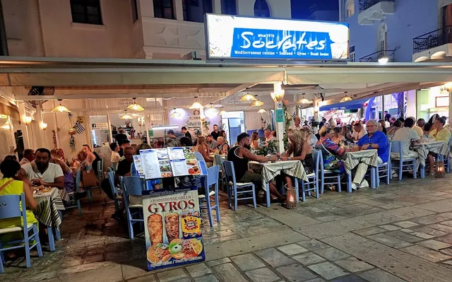 A busy taverna in the evening with pavement tables, checked tablecloths and a lit sign above