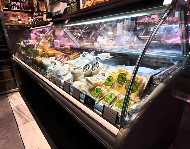 A refrigerated display case loaded with cheeses and cured sausages, with bottle shelves above it