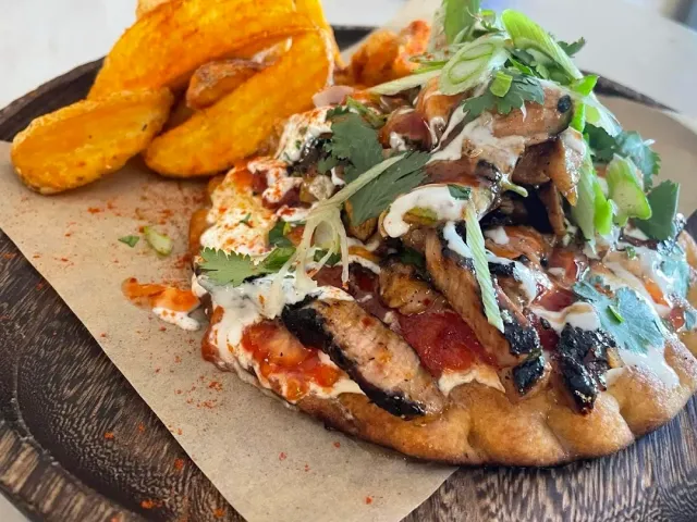 A flatbread loaded with grilled chicken pieces, sauces and coriander, beside potato wedges