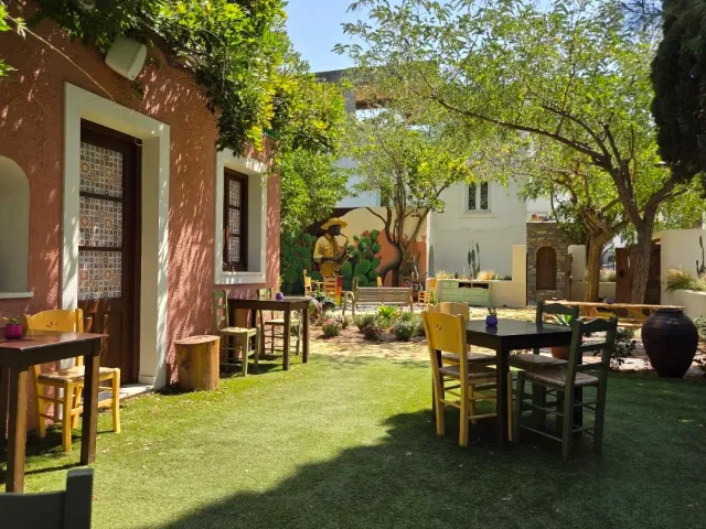 A lawn garden with tables and yellow chairs, a pinkish building and a mural on a white wall