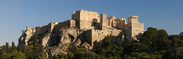 A rocky hill with ancient walls and marble temples on top, framed by pine trees