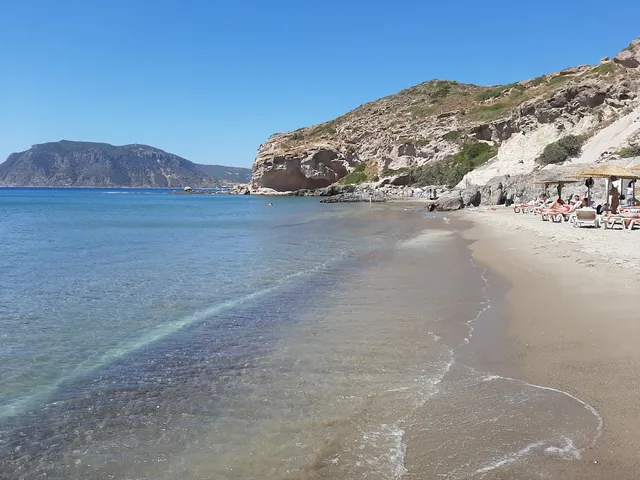 A sandy beach with clear water, sun loungers and straw umbrellas at the foot of a cliff