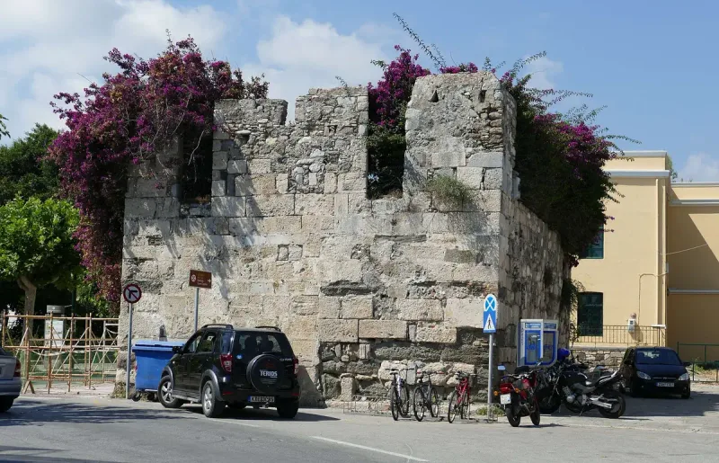 A bougainvillea covered medieval stone wall remnant in Kos old town, with a street and parked cars in front