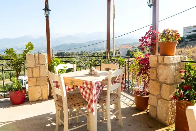 A covered terrace with a red checked table, flowering pots and a view of a valley and mountains