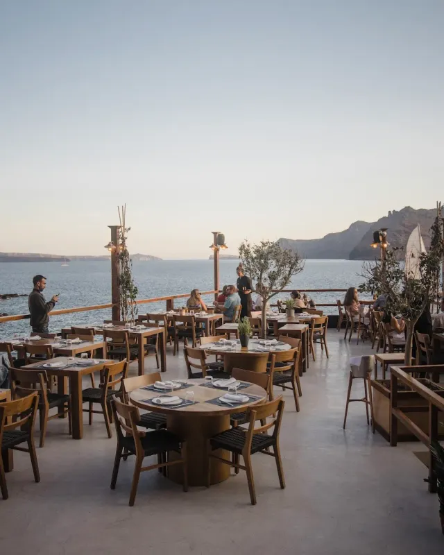 A wide terrace with round wooden tables, facing a bay and cliffs at dusk