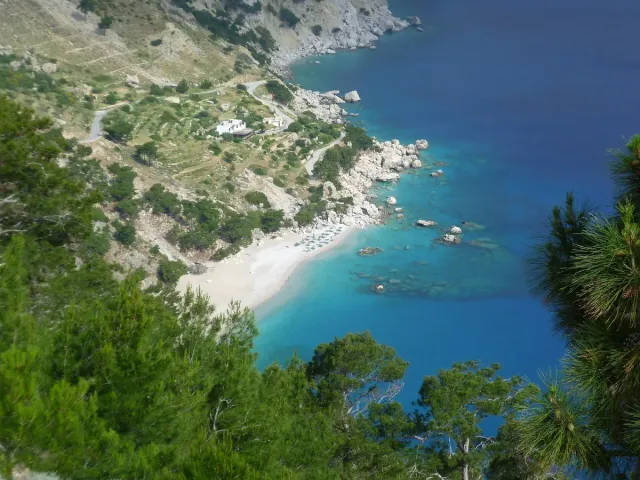A bay of clear blue water between cliffs and pine trees, with a pale pebble shore