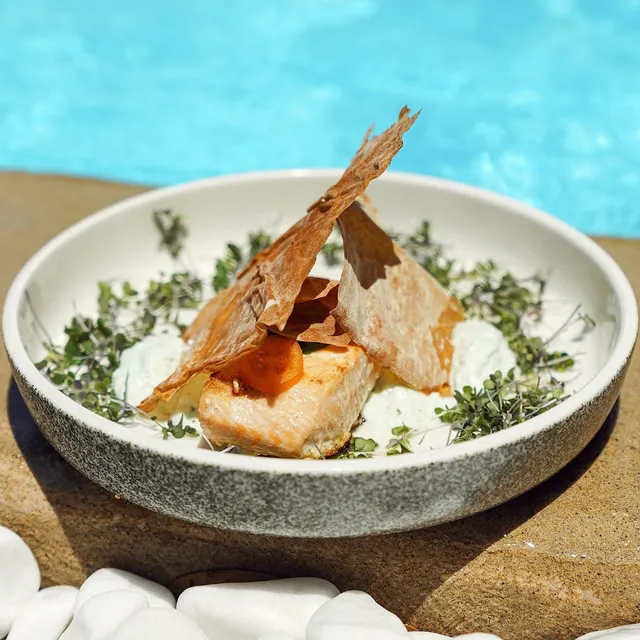 A fish dish with crispy pastry leaves and sprouts in a bowl, beside blue pool water