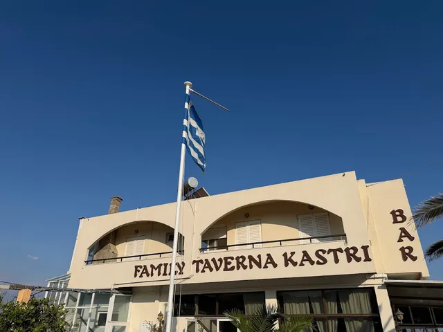 A pale building with arches and a Greek flag flying above it, under blue skies