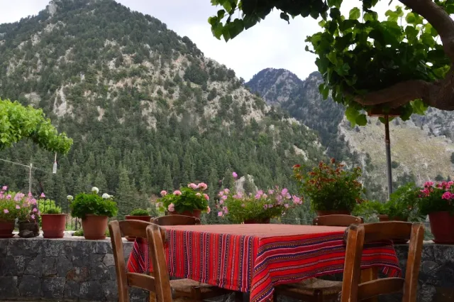 A table with a red woven cloth and wooden chairs, geranium pots on a wall facing forested mountains
