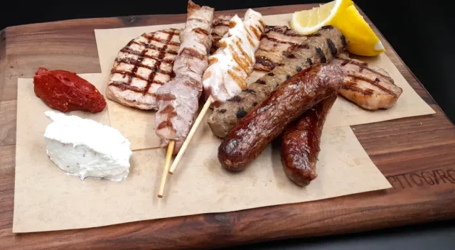 A wooden board with grilled meat cuts, skewers, sausages and two dips