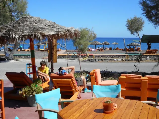 A terrace with a thatched parasol, orange wooden armchairs and turquoise fabric chairs facing the sea