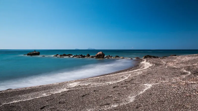A pebble shoreline with scattered rocks in the water and a ridge on the horizon
