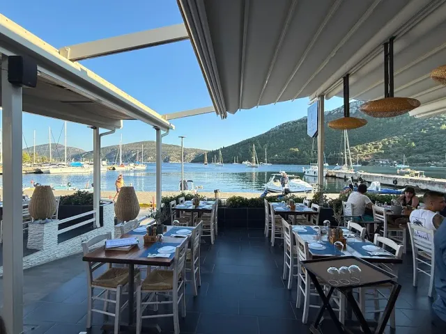 A covered terrace with wooden tables, facing a turquoise bay with moored yachts