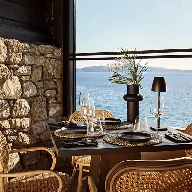 A table laid with black plates and wine glasses, wicker chairs and a stone wall facing a window to the sea