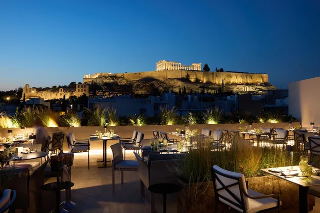 A rooftop lit up in the evening with set tables, and the illuminated Acropolis in the background