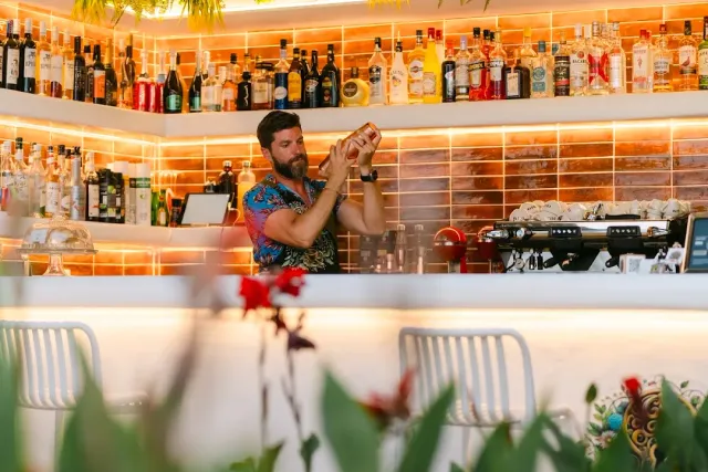 A bartender shaking a shaker behind a lit counter with shelves of bottles, and blurred plants in front