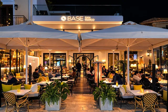 Tables full of diners under large white parasols, in front of a lit facade in the evening
