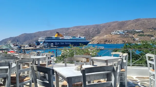 A terrace with grey wooden tables and chairs facing a bay with a large ferry and hills