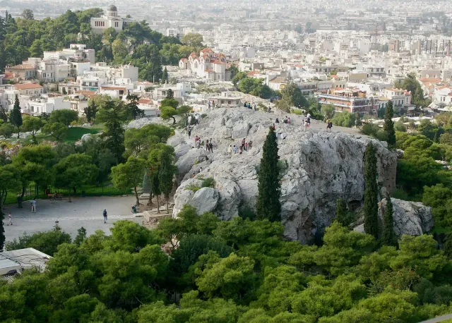 A bare marble rock with visitors climbing it, the city spreading behind to the horizon