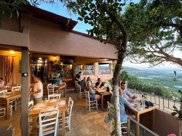A tree shaded terrace with tables and diners, and an open view of a plain and the sea toward evening