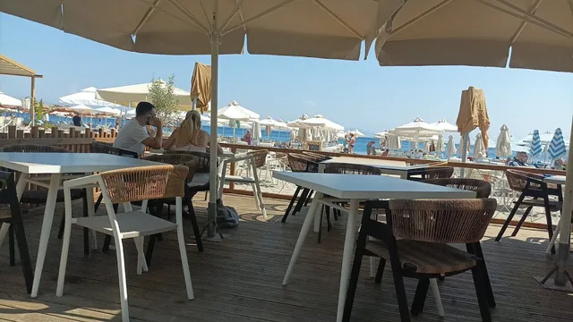 White tables and woven chairs on a wooden deck under umbrellas, facing a busy beach
