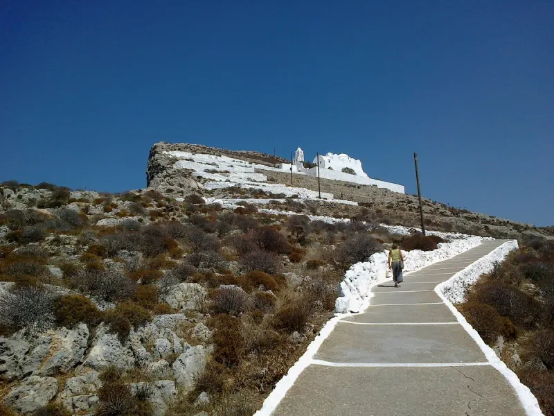 A whitewashed path zigzagging up a rocky slope to a white complex on the ridge, with a lone walker climbing
