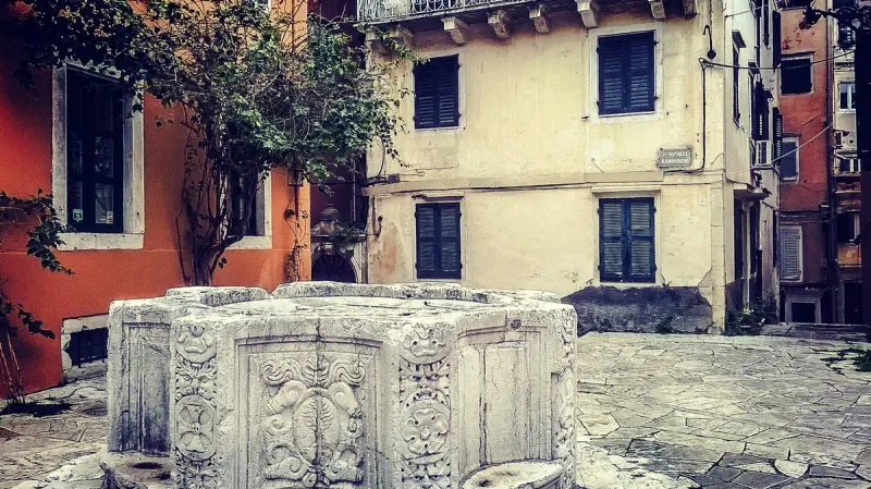 A carved stone wellhead in a small square between butter and terracotta coloured houses
