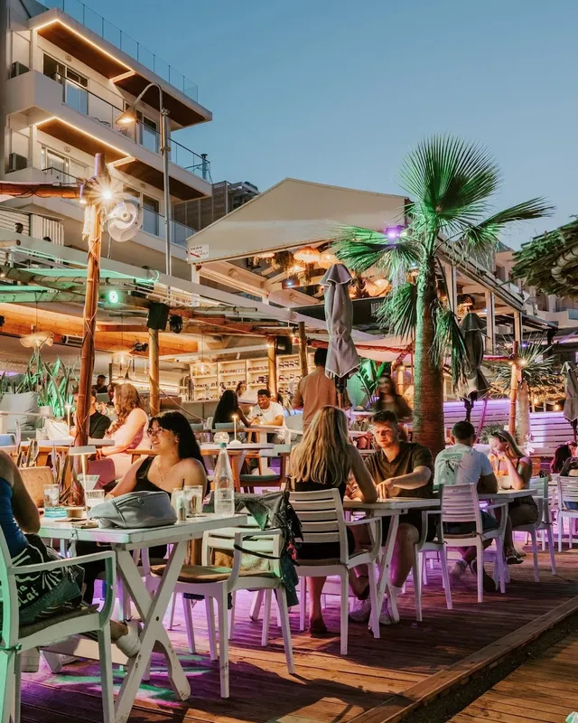 People sitting at white tables on a wooden deck at dusk, under a palm tree and lights