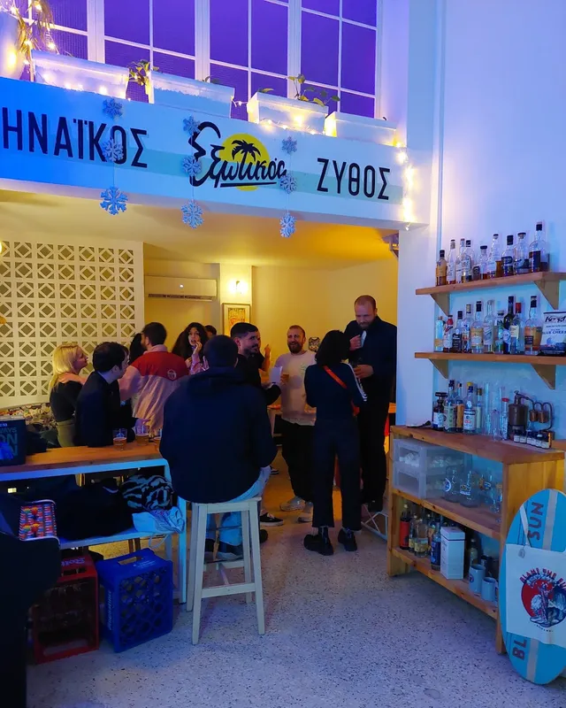 A small bar lit up in the evening, people standing with beers and bottle shelves along a wall